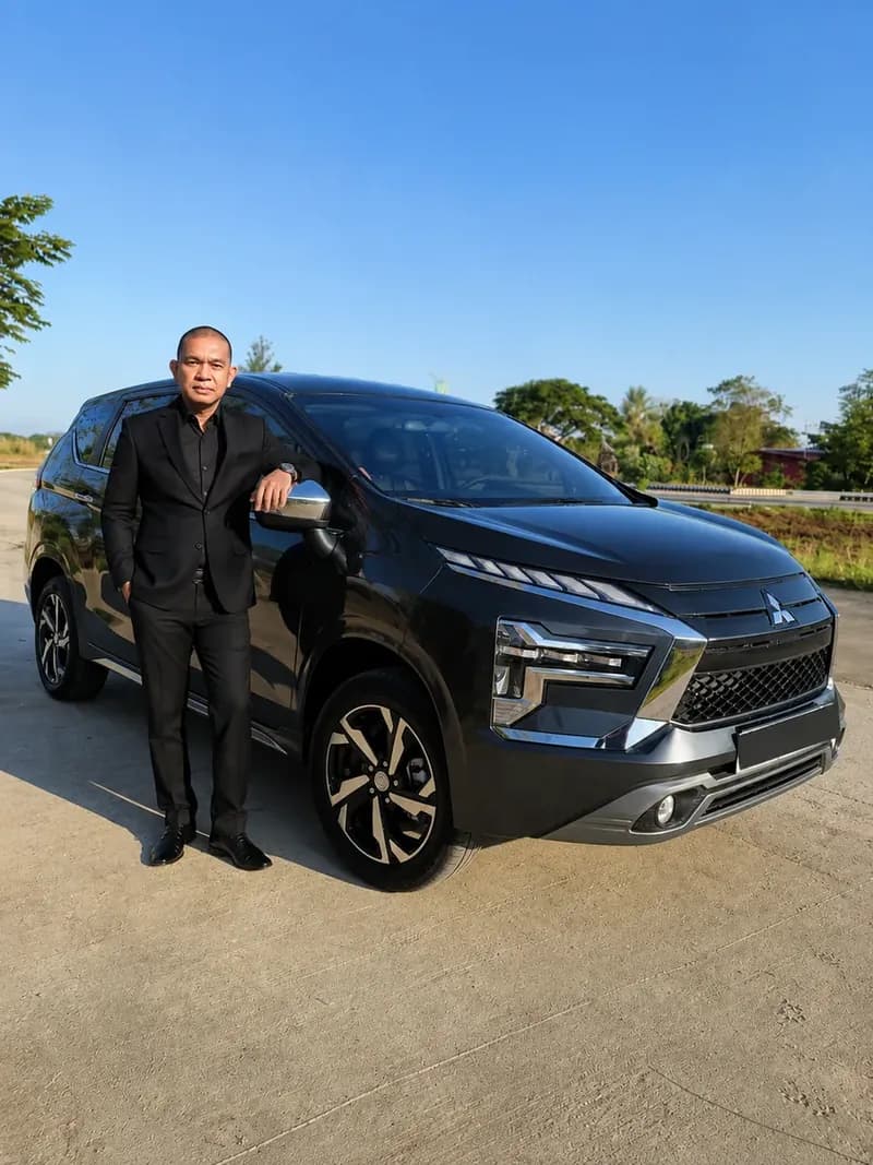 Cris Labastida in a black suit, standing beside the graphite Mitsubishi Xpander on an open road